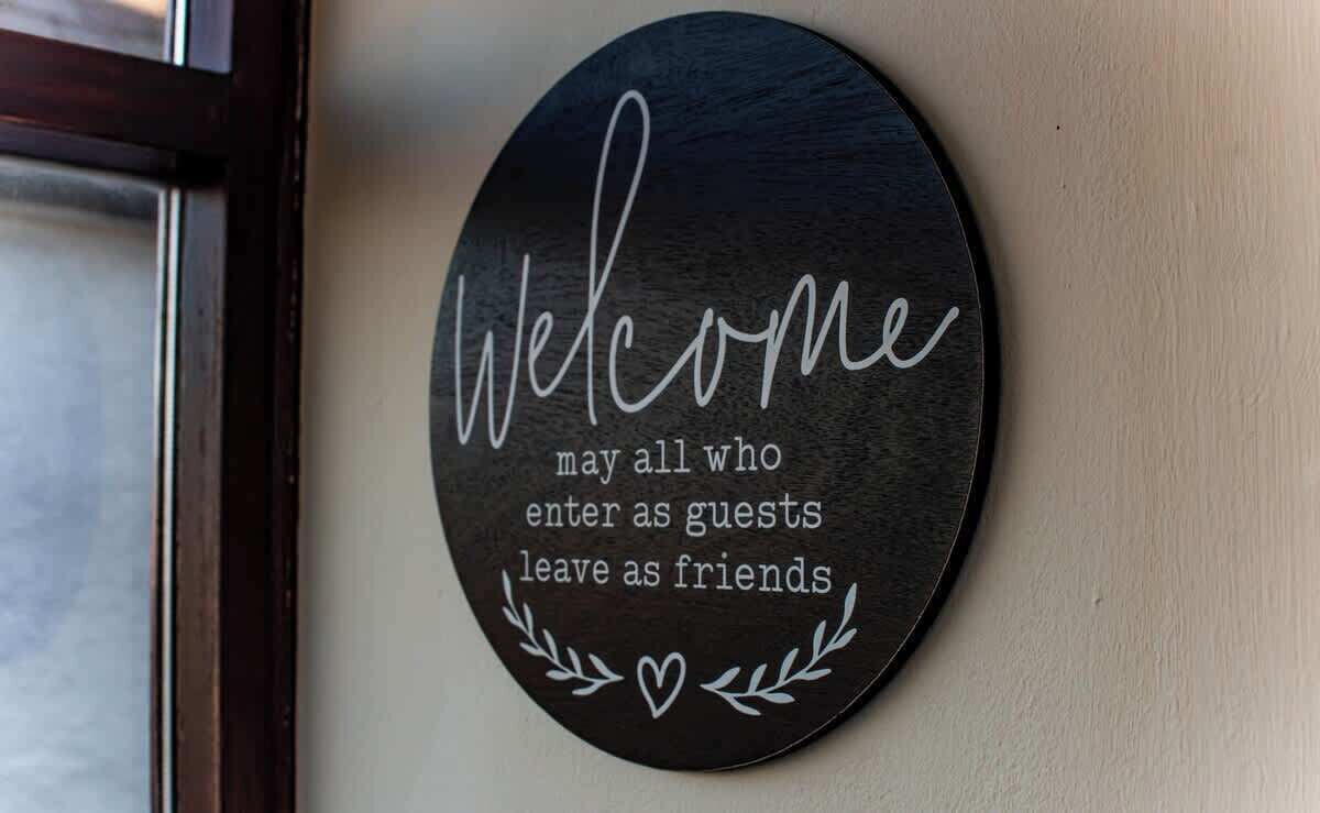 Een rond bord met de tekst 'Welcome may all who enter as guests leave as friends' op een muur.