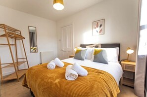 Hotellihuone, jossa on sänky, puinen pyyheteline, yöpöytä lampun kanssa ja seinällä oleva kehystetty taideteos.