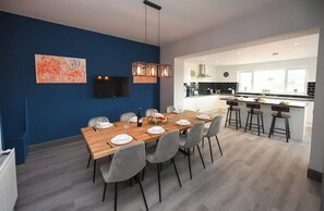 A modern dining area with a wooden table set for six, grey chairs, and a kitchen in the background.