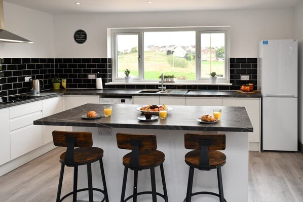 Una cocina moderna con una isla central, tres taburetes, un refrigerador y un ventanal con vistas al exterior.