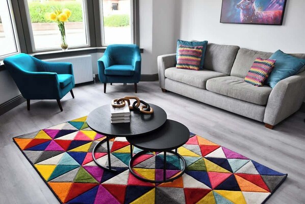 A living room with a grey sofa, two blue armchairs, a black coffee table with books, and a colorful geometric rug.