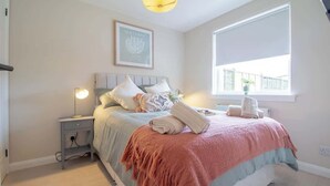 A bedroom with a bed, bedside table, lamp, window, and artwork on the wall.