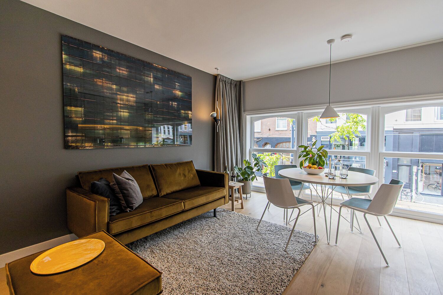 Een moderne woonkamer met een bank, een eettafel met stoelen, een groot raam met gordijnen en uitzicht op de stad.