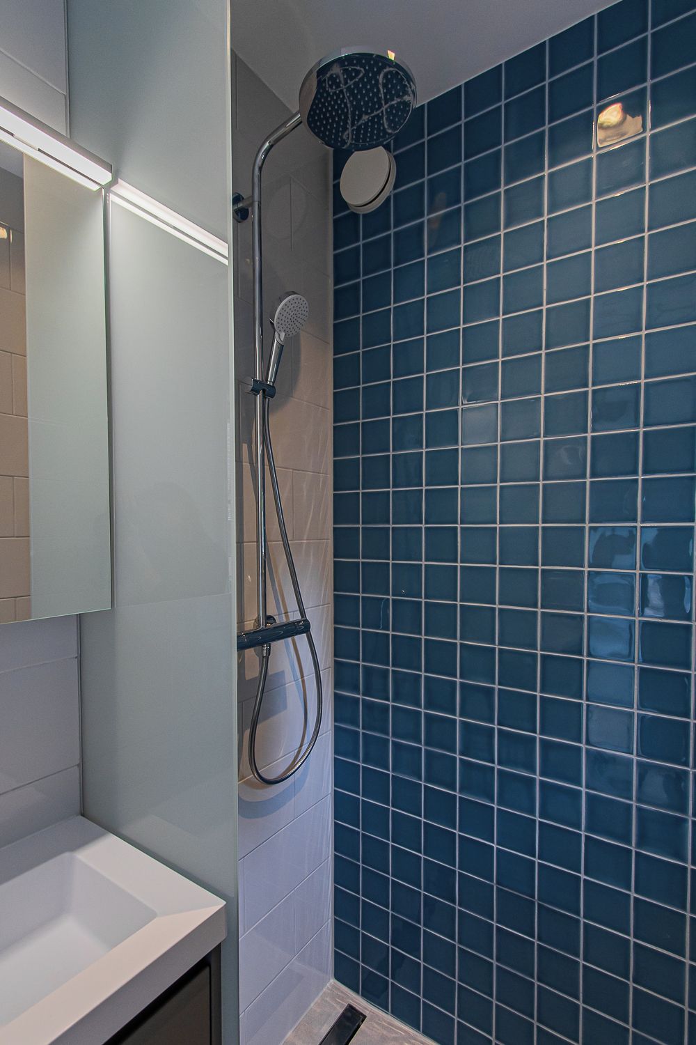 Une salle de bain avec un mur recouvert de carreaux bleus, un lavabo blanc et une douche équipée d’un pommeau de douche à main.
