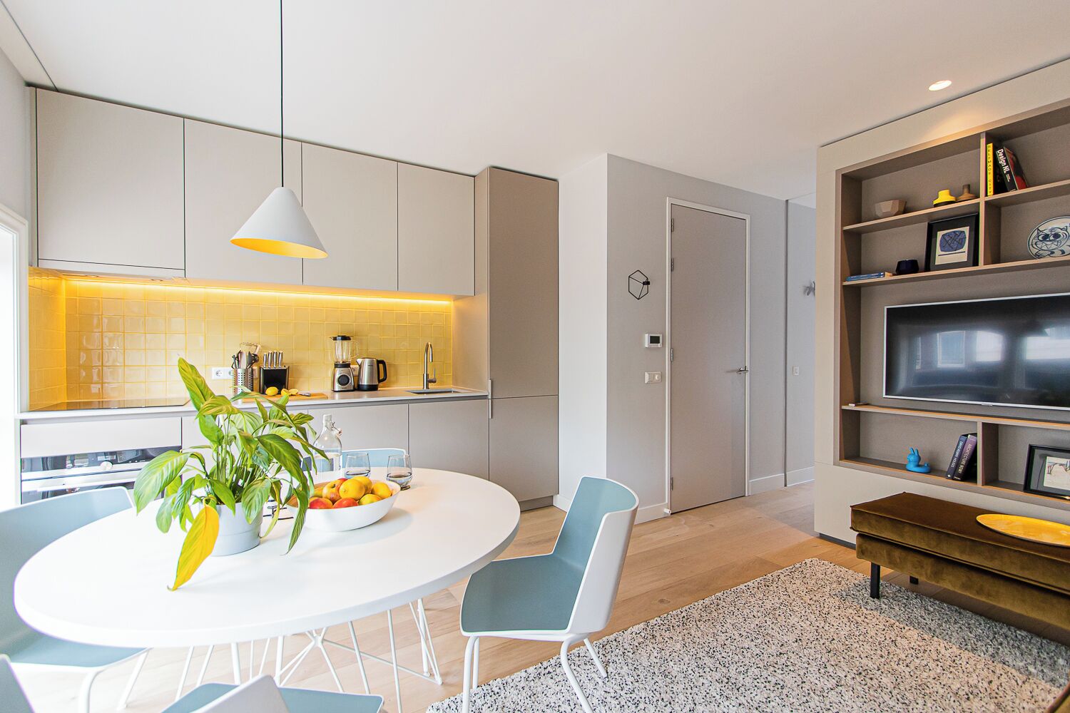 A modern kitchen with a dining area, including a white table with chairs, a potted plant, and a bowl of fruit.