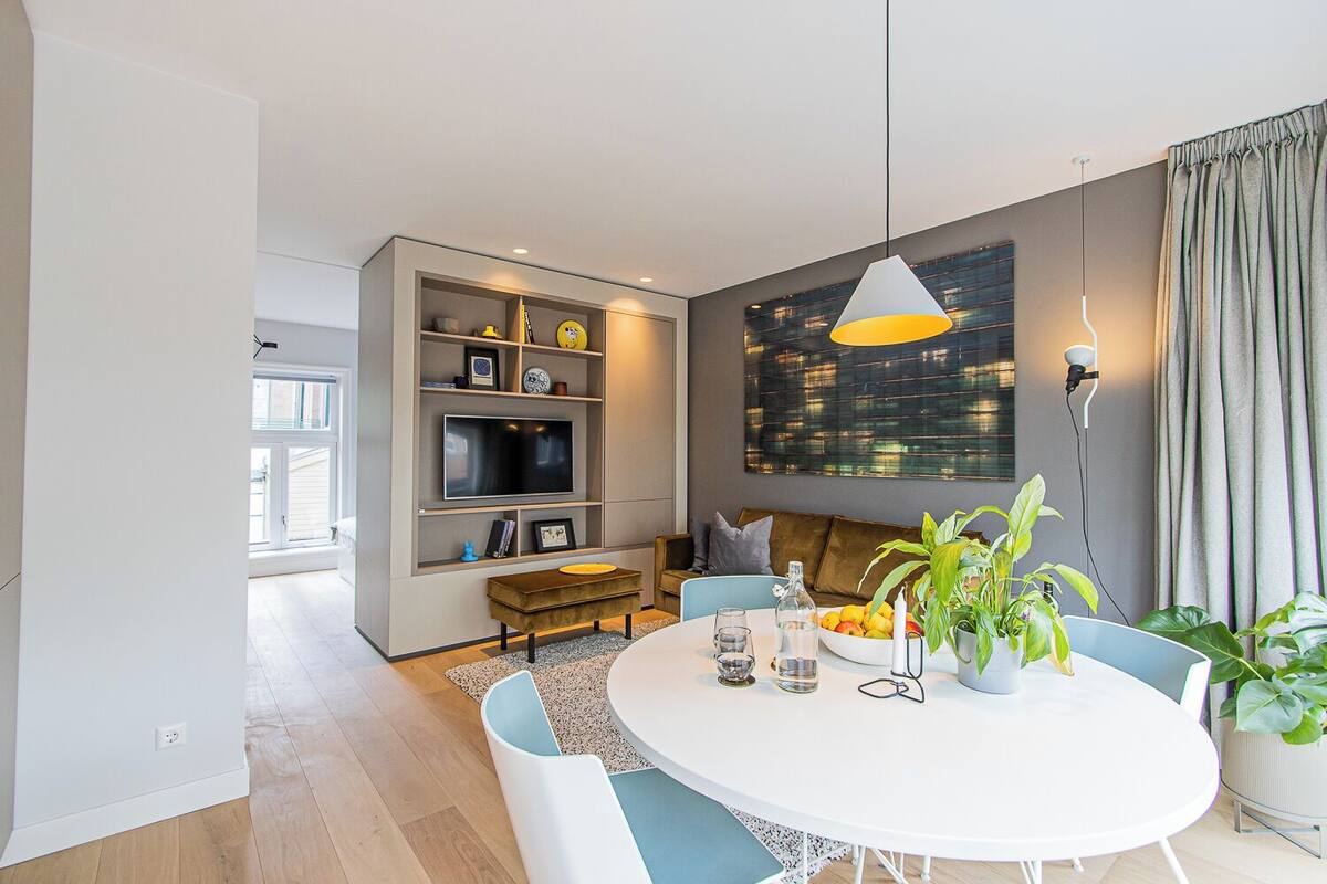 A modern living room with a white round table, a sofa, and a TV mounted on a wall. There is a large window with a view of the city, and a plant on the right side.