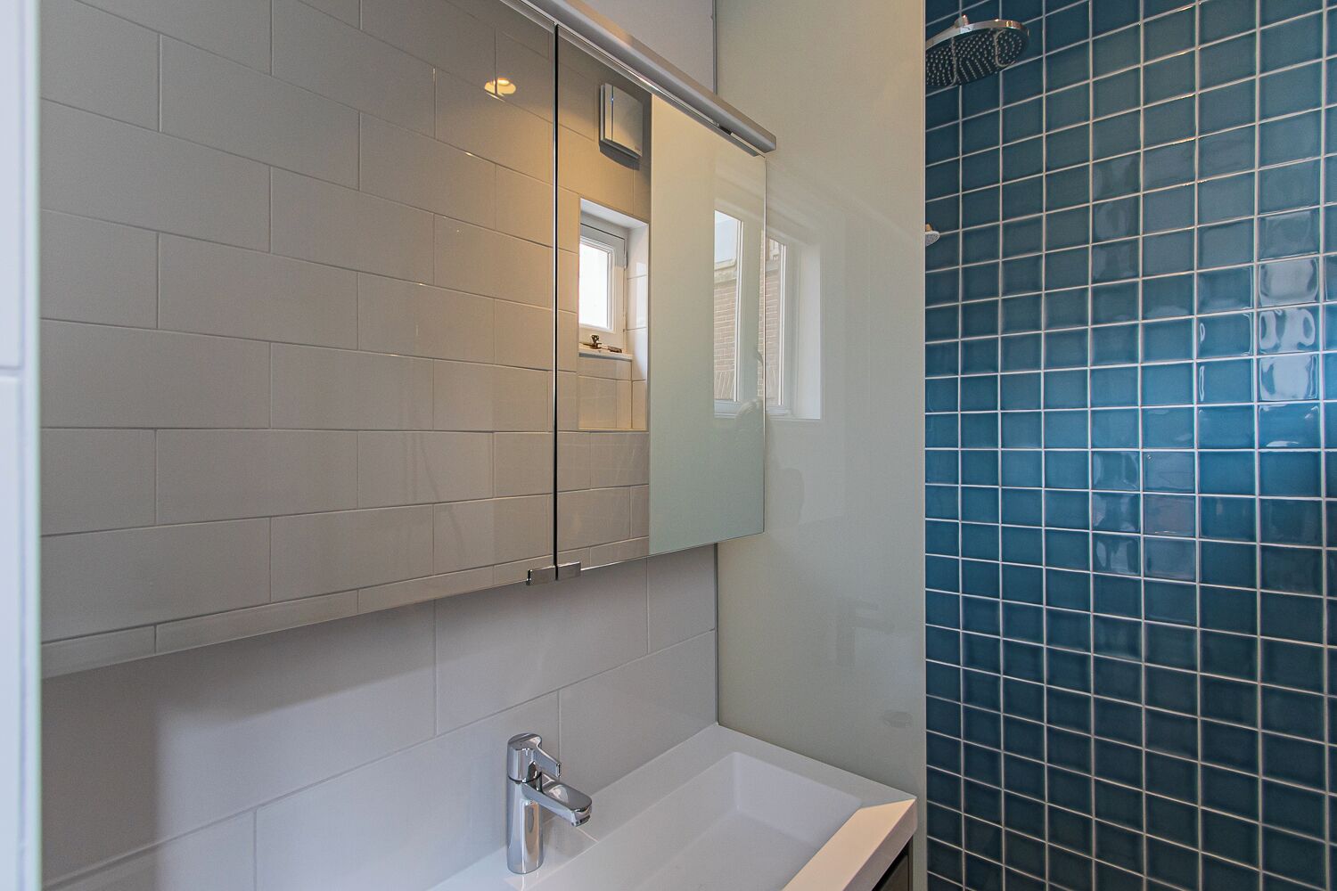 A bathroom with white tiles, a mirror, a sink, and a shower area with blue tiles.