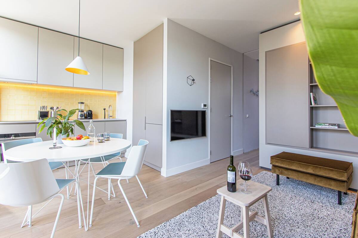 A modern kitchen with white cabinets, a dining area with a round table and chairs, a TV mounted on the wall, and a bench in the living area.