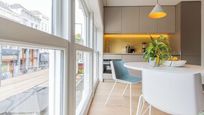Moderni keittiö, jossa on suuret ikkunat, ruokailutila pöytineen ja tuoleineen sekä näkymä ulos kadulle.