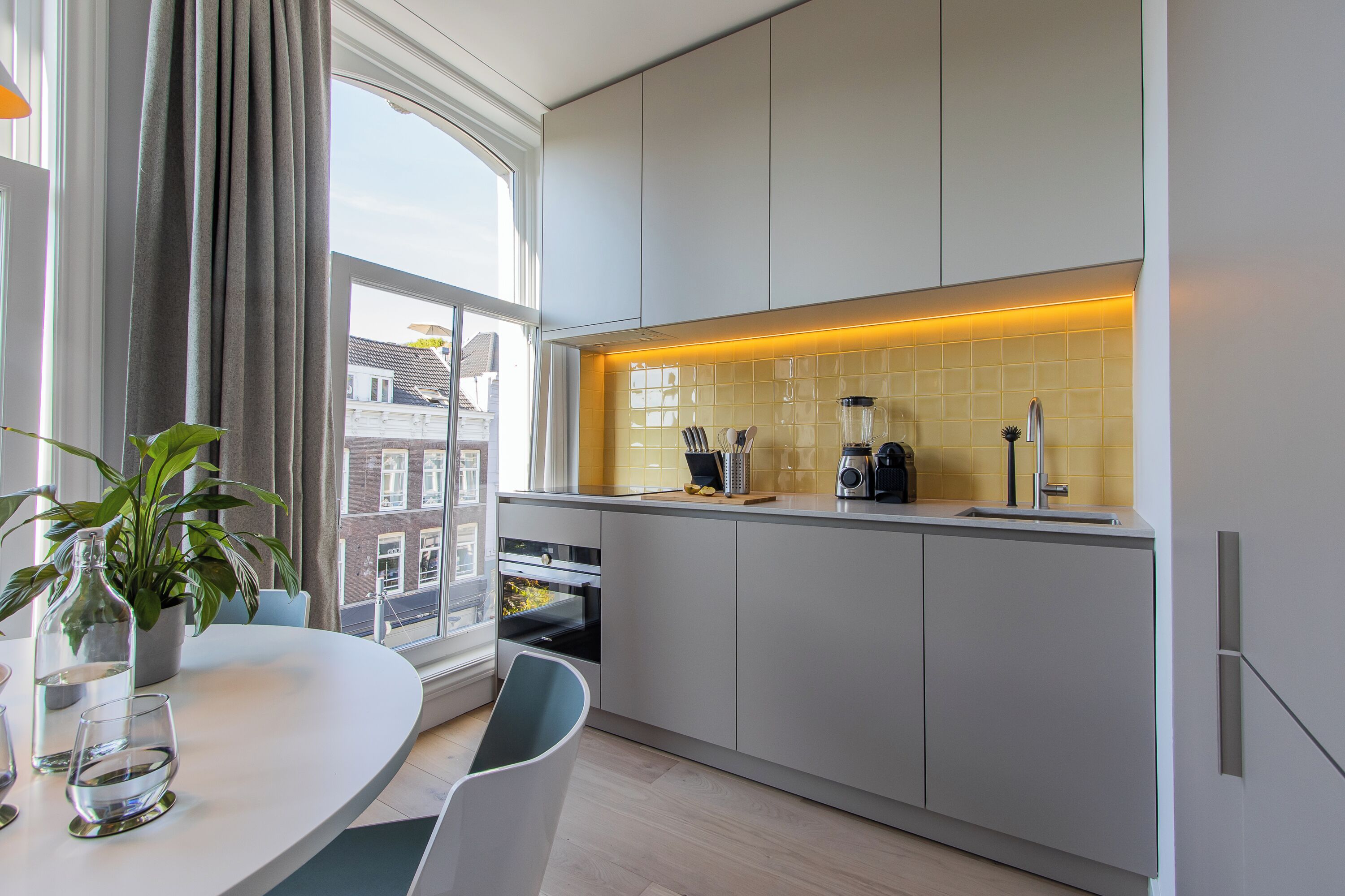 A modern kitchen with white cabinets, a built-in oven, a coffee machine, and a window with curtains.