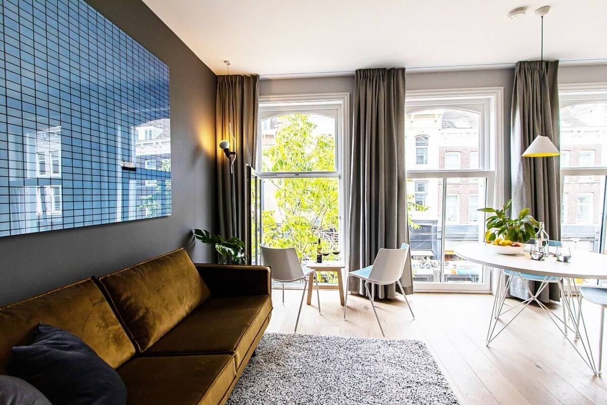 A modern living room with a brown sofa, a dining table with chairs, and a large window with curtains.