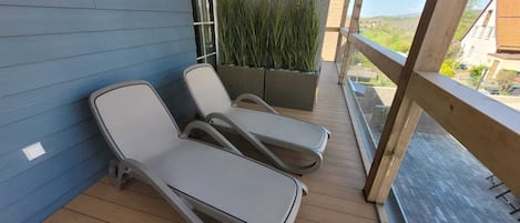 Un balcon avec deux fauteuils de jardin, une terrasse en bois, un mur bleu et une vue sur un quartier résidentiel.