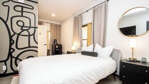 Habitación de hotel con una cama grande, una mesita de noche con lámpara, un espejo y una pared decorada con arte abstracto.