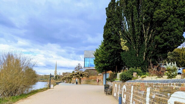 A paved pathway alongside a river, with a brick wall on one side and a modern building on the other.