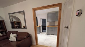 A living room with a brown leather sofa, a wooden-framed door leading to a kitchen with grey cabinets and a white tiled floor.
