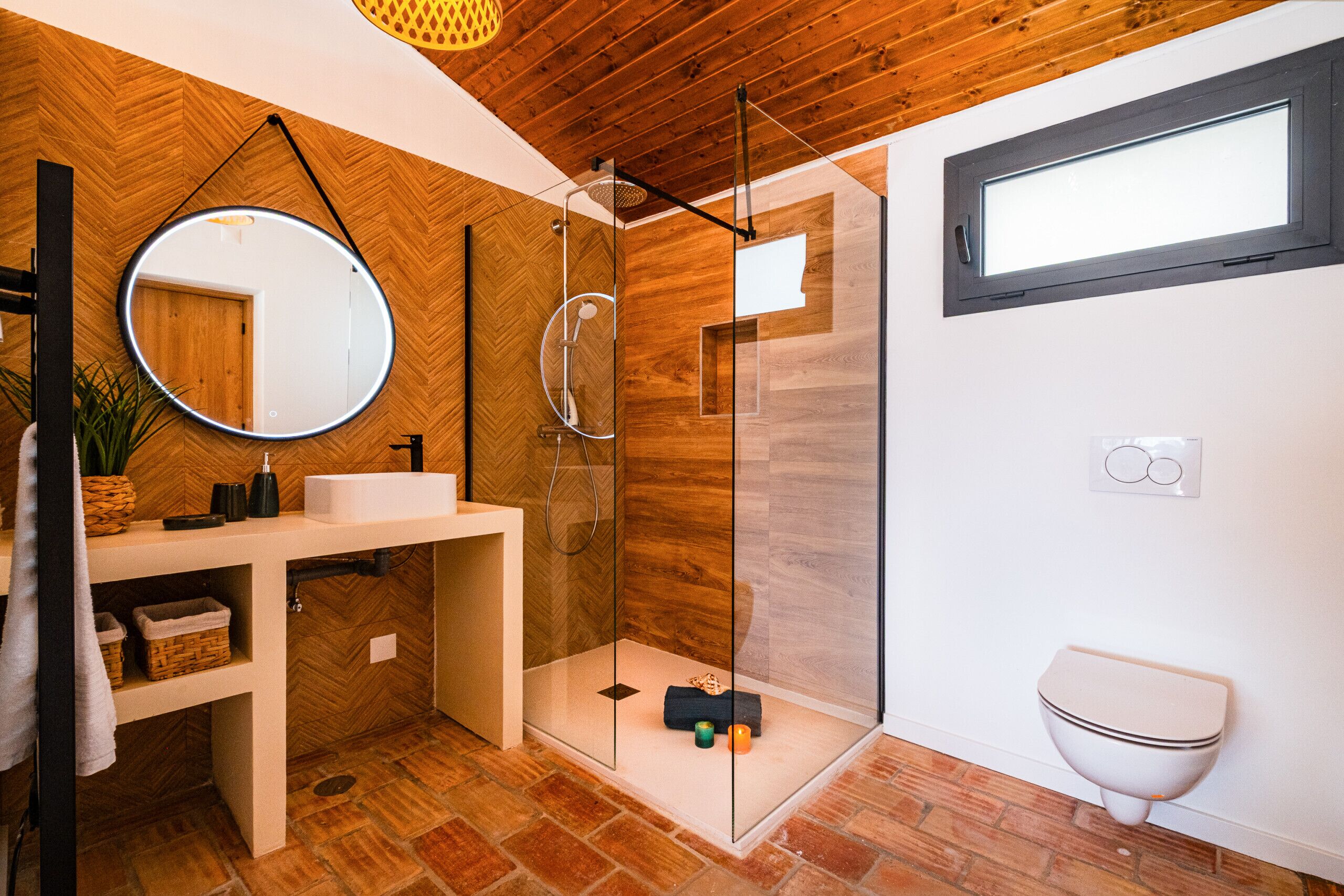 A modern bathroom with a wooden herringbone floor, a round mirror, a glass-enclosed shower, and a toilet.