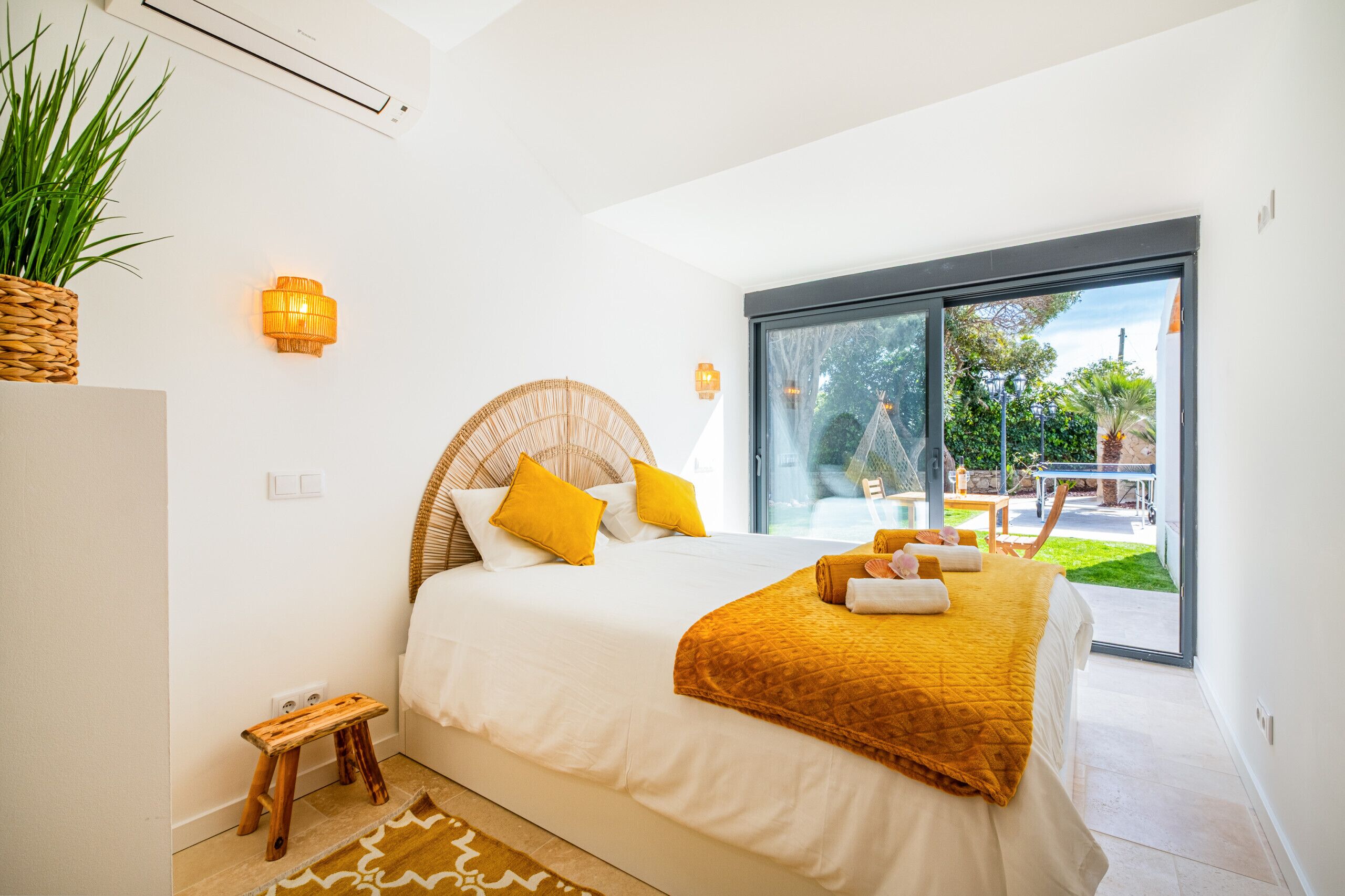 A bedroom with a large bed, a small wooden stool, a potted plant, and a view of an outdoor area with a pool and patio.