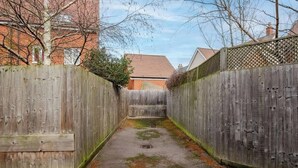 A narrow alleyway with a wooden fence on both sides, a number '48' painted on the ground, and a building with a red roof in the background. - Spacious 4 Bed Home With 2 Bathrooms & Parking (Bedford)