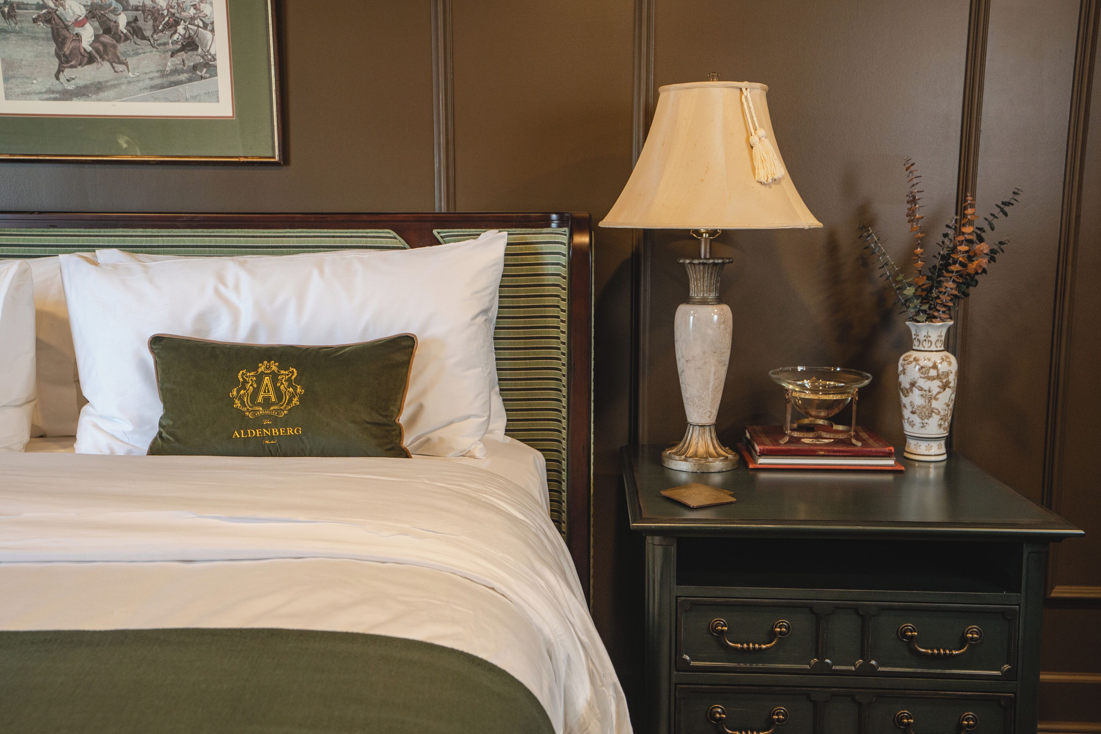 A neatly made bed with a green pillow and a white pillow, a bedside table with a lamp, a vase with flowers, and a framed picture on the wall.