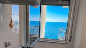 Una habitación con vistas al mar a través de un ventanal grande, un tendedero metálico y una pared blanca con un enchufe.