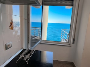 A room with a view of the sea through a large window, a metal drying rack, and a white wall with a power outlet.