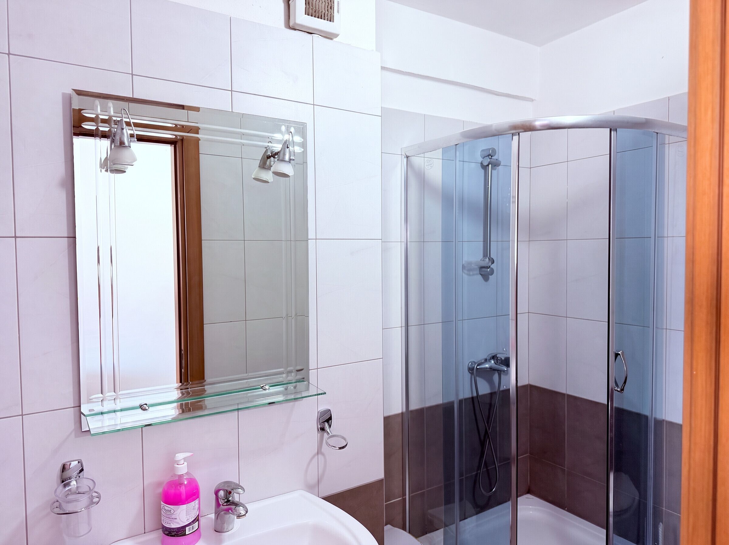 A bathroom with a glass shower enclosure, a mirror with a glass shelf, and a white sink with a soap dispenser.