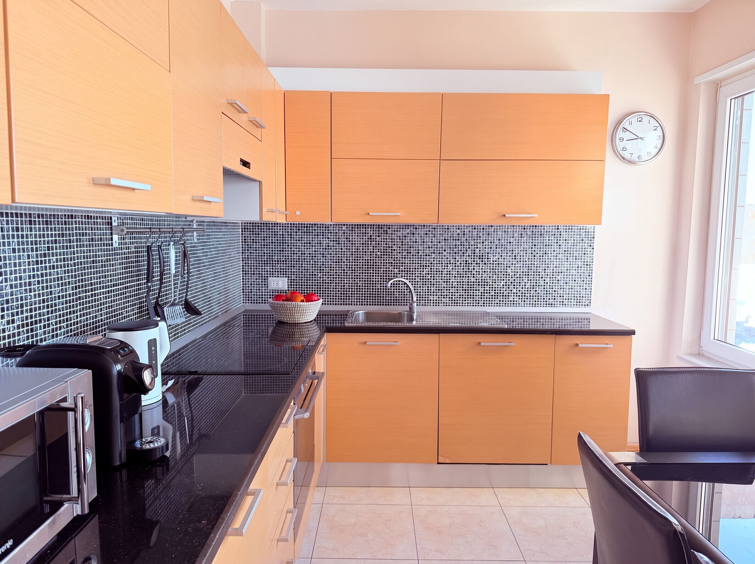 A modern kitchen with black countertops, wooden cabinets, a tiled backsplash, a coffee machine, and a bowl of fruit.