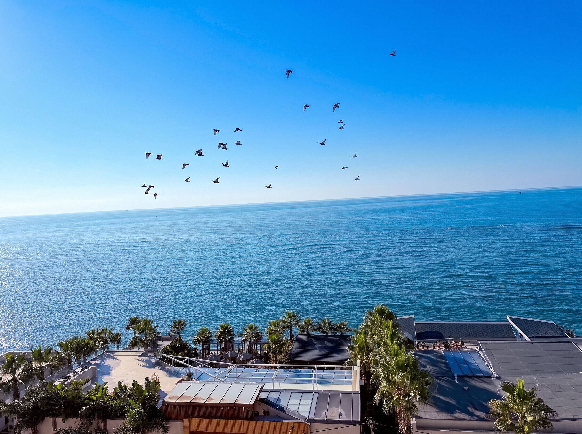 A coastal view with a clear sky, a beach, and a building with a rooftop terrace.