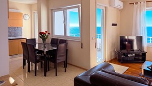 A living room with a sofa, a TV, a dining area with chairs and a table, a kitchen with cabinets, a clock, and a view of the sea through a window.