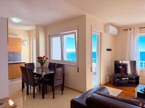 Ein Wohnzimmer mit einer Couch, einem Fernseher, einem Essbereich mit Stühlen und einem Tisch, einer Küche mit Schränken, einer Uhr und Meerblick durch ein Fenster.
