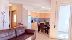 A modern living room with a leather sofa, a dining area with a table and chairs, and a kitchen with wooden cabinets and a refrigerator.