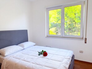 Ein ordentlich bezogenes Bett mit weißer Bettwäsche, eine einzelne rote Rose, ein grauer Kopfteil und ein Fenster mit Blick auf grüne Bäume.