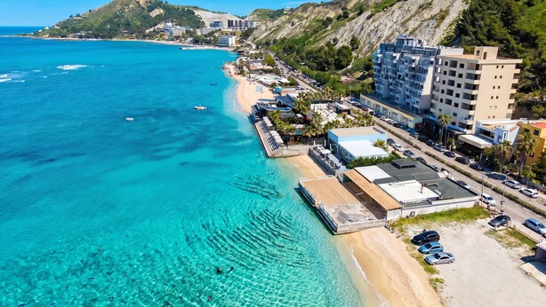 A coastal hotel with a beach view, a parking area, and a hillside backdrop.