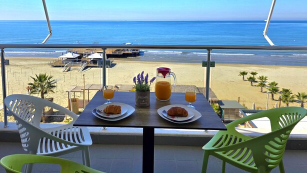 Un balcón con una mesa dispuesta para el desayuno, con vista a una playa con palmeras y un cielo azul despejado.