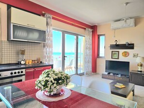 Une cuisine moderne équipée d’une cuisinière Zanker, une table à manger en verre agrémentée d’un centre de table floral blanc, un téléviseur et un balcon offrant une vue sur la mer.