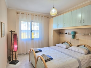 Un dormitorio con cama, lámpara de noche, armario y ventana con cortinas.