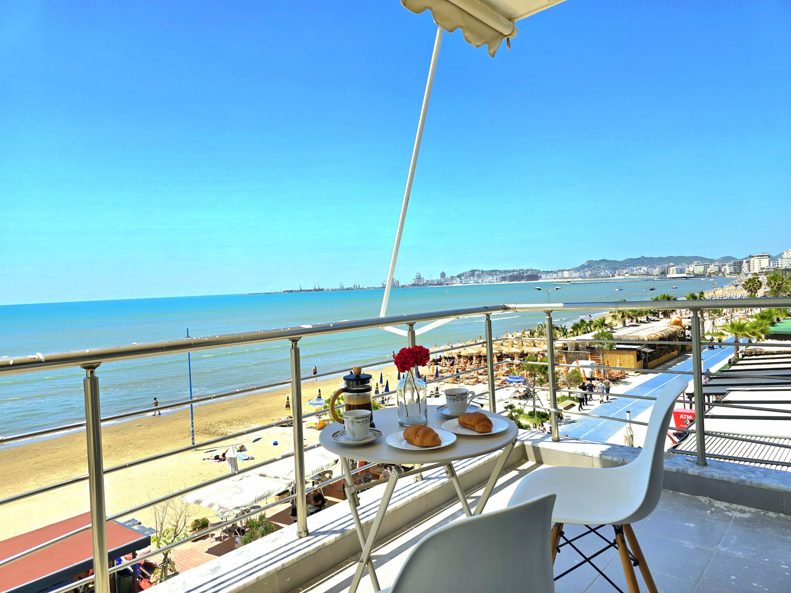 A balcony with a table set for breakfast, overlooking a beach with umbrellas and people.