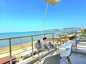 Un balcon avec une table préparée pour le petit-déjeuner, donnant sur une plage parsemée de parasols et animée par la présence de nombreuses personnes.