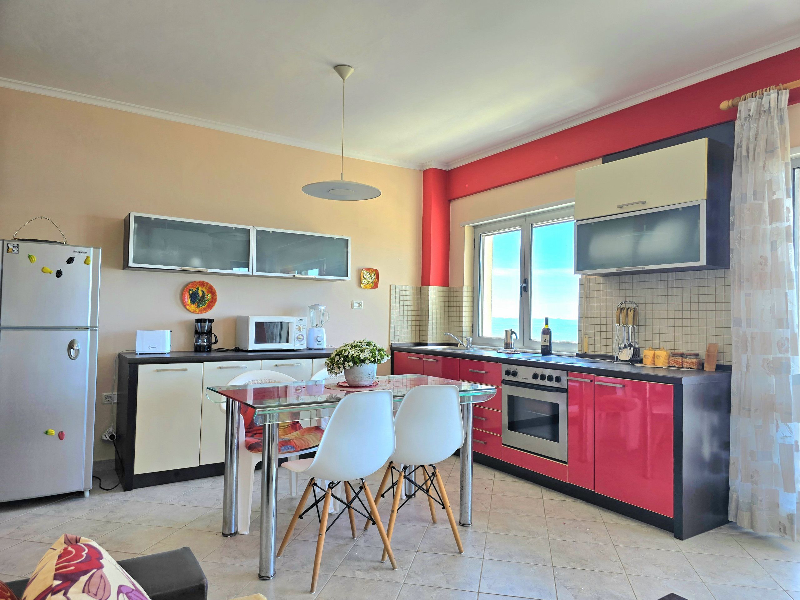A modern kitchen with a red and beige color scheme, featuring a dining table with chairs, a refrigerator, a microwave, a stove, and a window with a view.