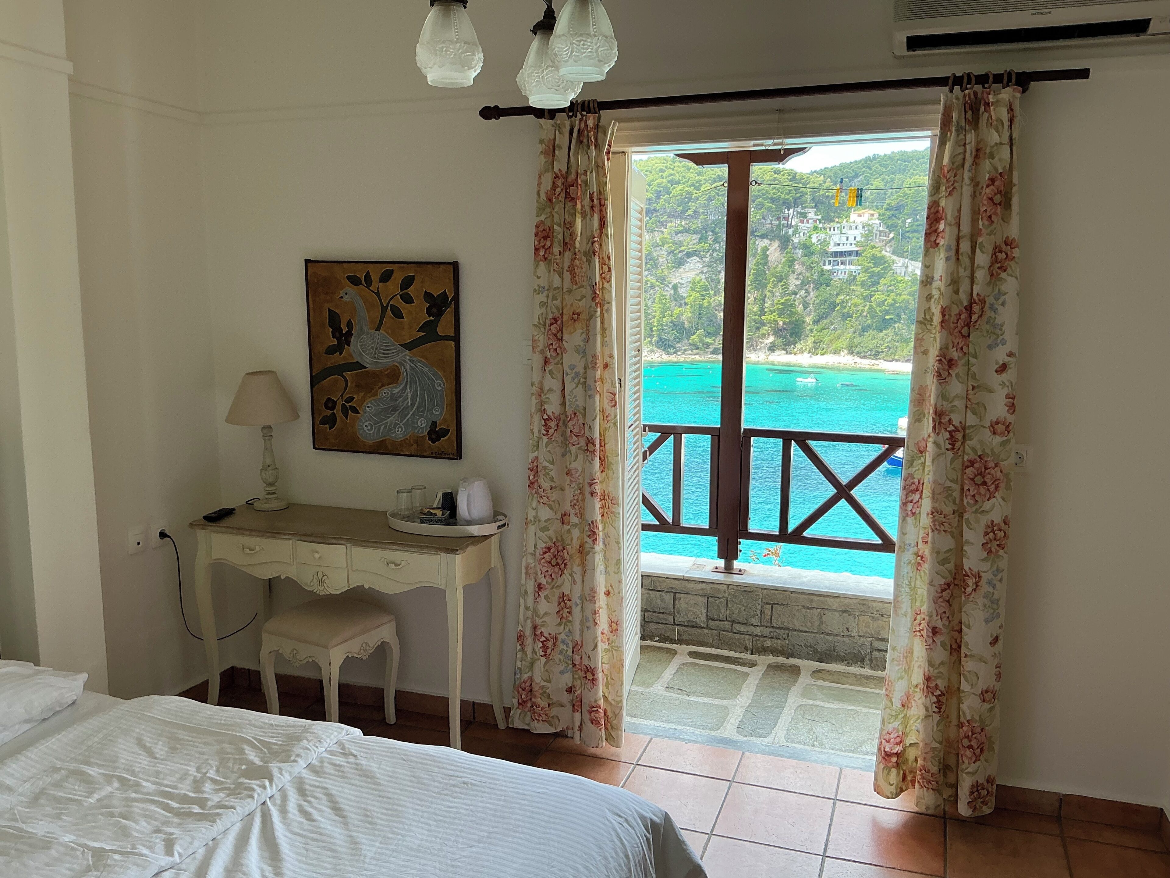 A bedroom with a bed, a desk with a lamp, a painting, and a view of a turquoise sea through curtains.