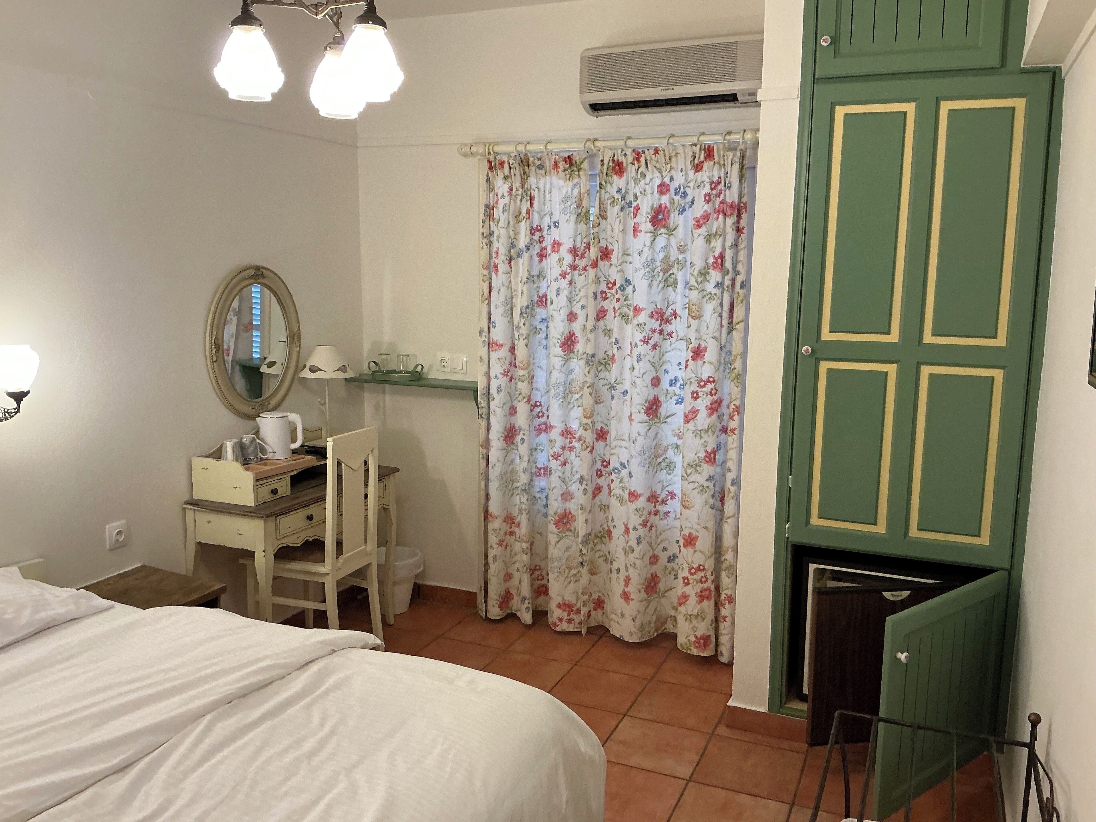 A bedroom with a bed, a desk with a chair, a mirror, a window with floral curtains, and a green door.