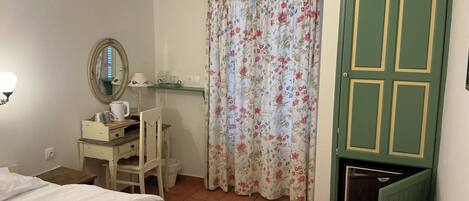 A bedroom with a bed, a desk with a chair, a mirror, a window with floral curtains, and a green door.