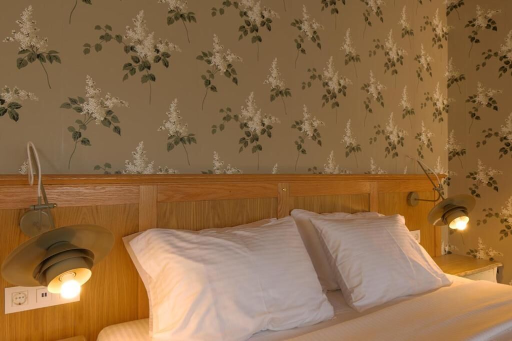A bedroom with a wooden headboard, two bedside lamps, and a floral wallpaper pattern.