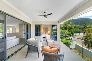 Un balcon moderne doté d’un ventilateur de plafond, de portes vitrées donnant sur un espace de vie avec un canapé et des fauteuils, et offrant une vue sur un paysage verdoyant luxuriant.