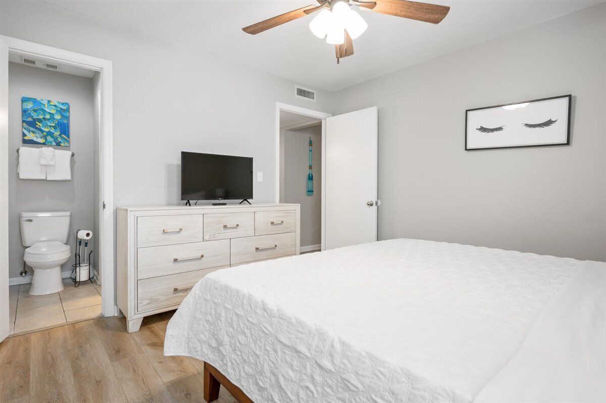 A bedroom with a white bed, a wooden dresser, a television, and a ceiling fan.