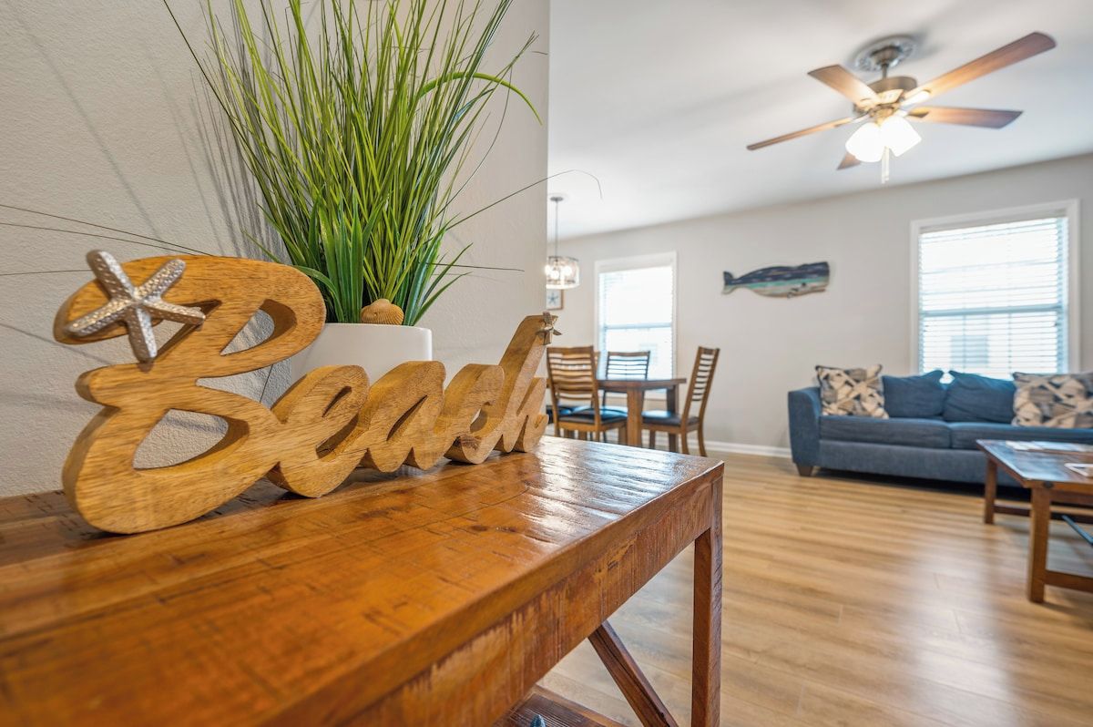A wooden table with a decorative word 'Beach' and a starfish ornament, a potted plant, and a ceiling fan in a living room with a blue sofa and a dining area.
