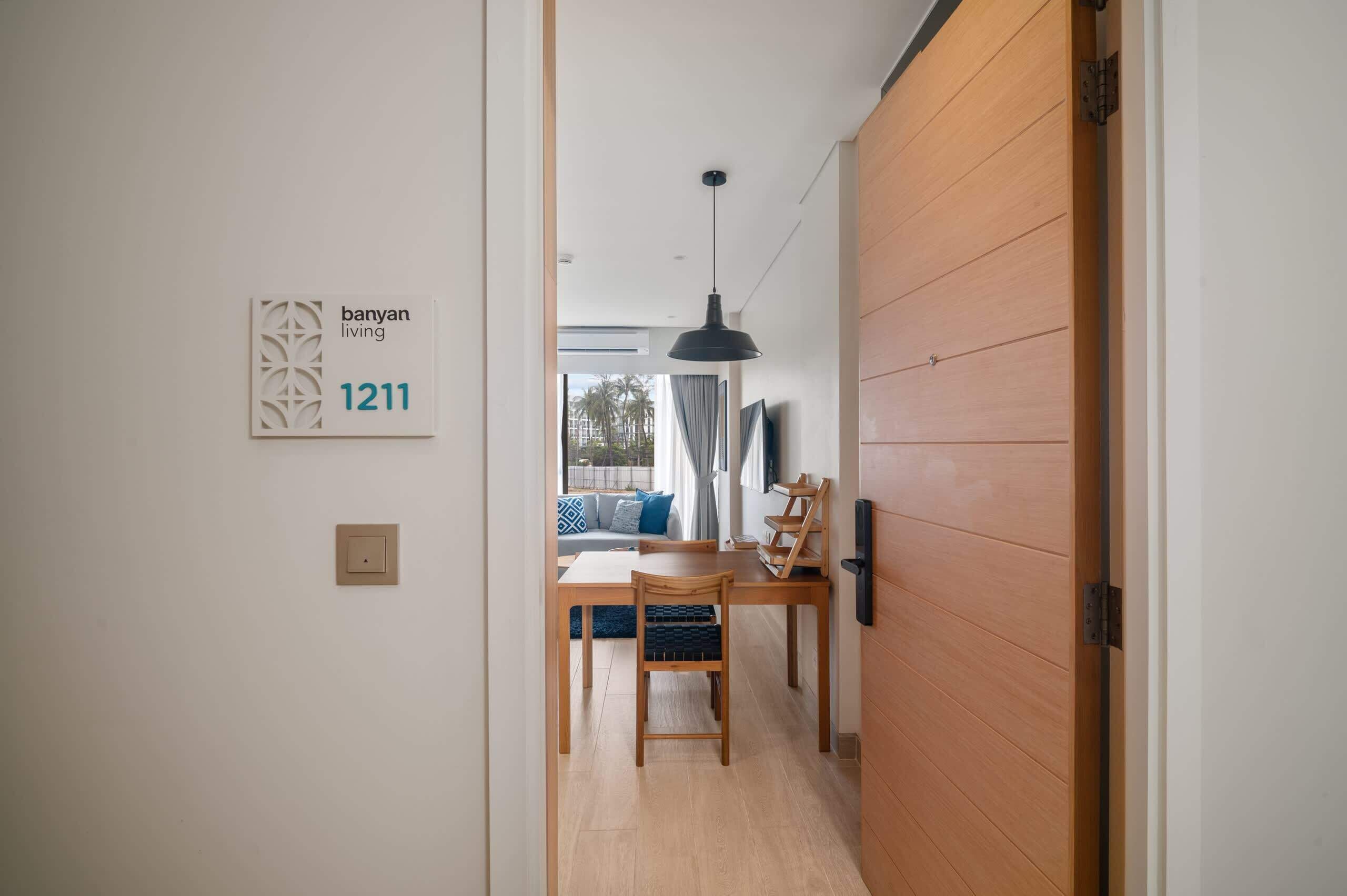 En hall leder till ett matområde med trägolv, ett matbord och en soffa. En hänglampa hänger från taket, och en skylt på väggen visar texten ”banyan living 1211”.