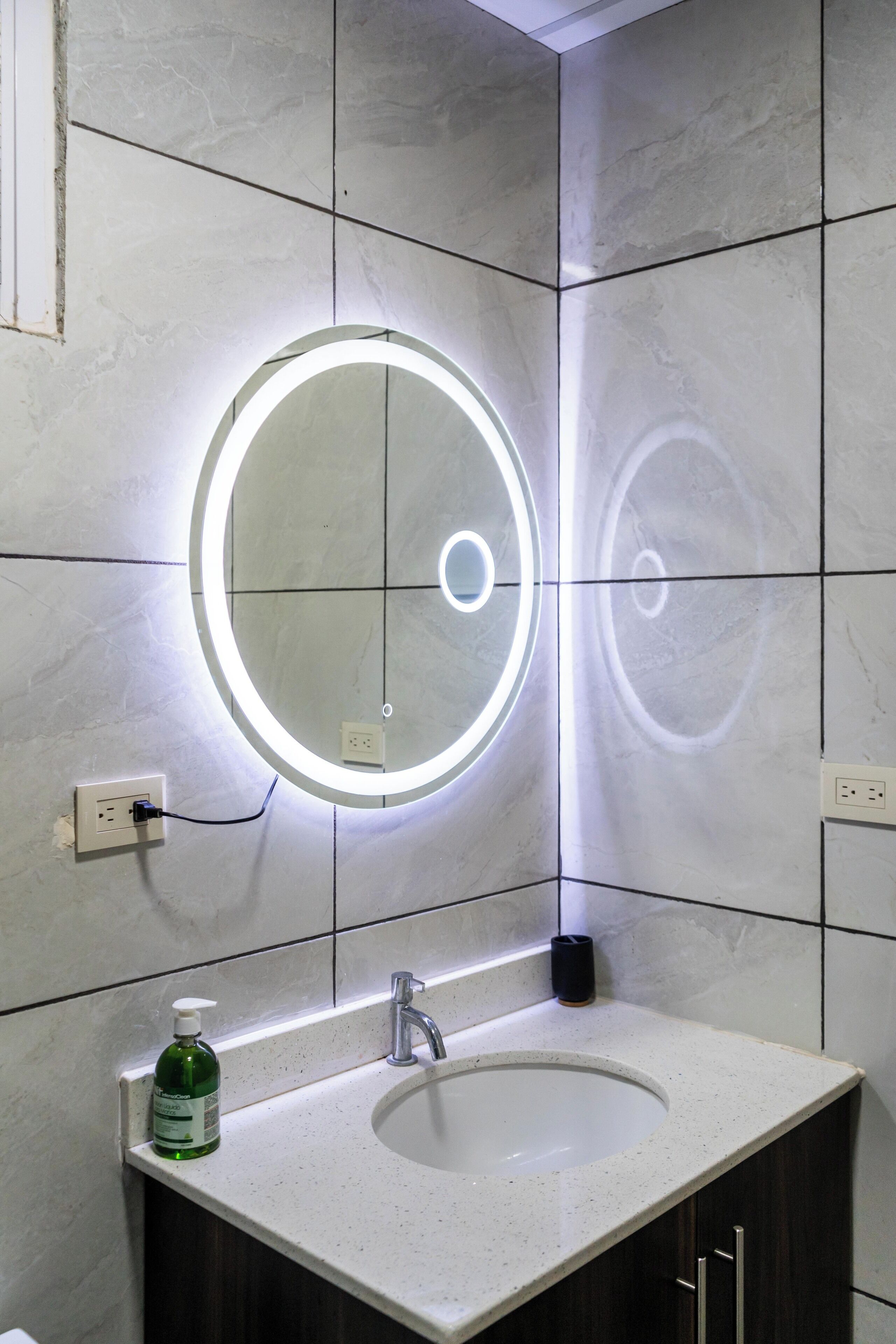 Un baño con un lavamanos blanco, un espejo con luz circular y una botella de jabón para manos.