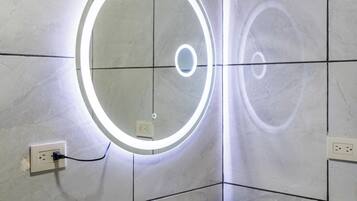 Un baño con un lavamanos blanco, un espejo con luz circular y una botella de jabón para manos.
