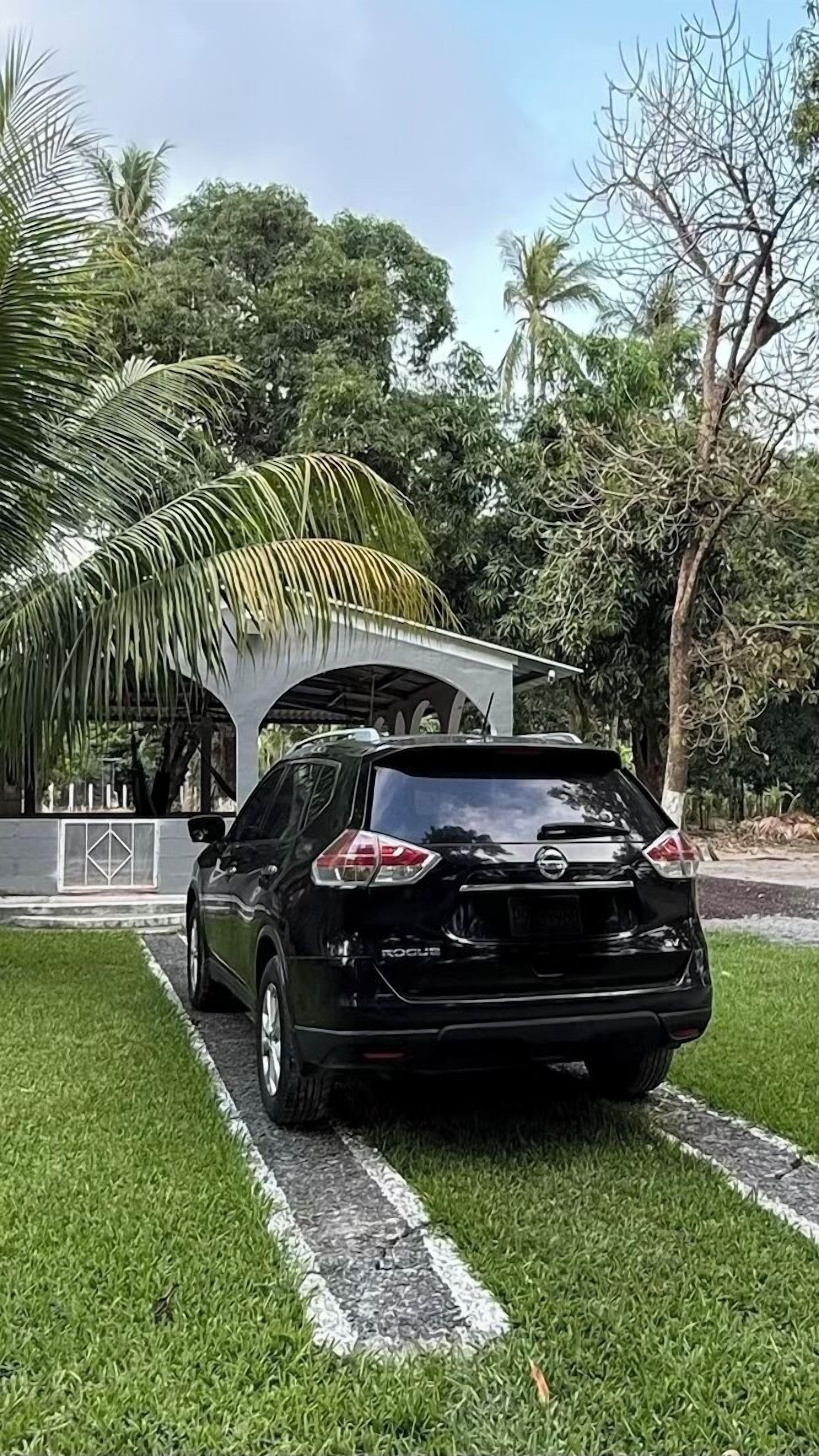 A black car parked on a grassy area with a covered structure and trees in the background.